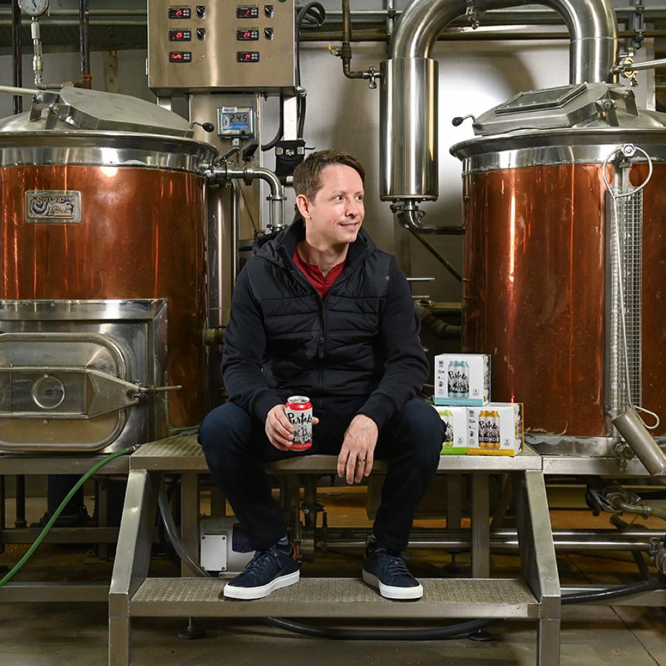 Ted Fleming, Sc’00, sitting on a bench in front of two distillers at his brewery.