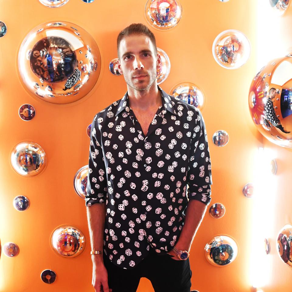 Simon Huck, in a black shirt with a dice pattern and black pants, standing in front of an orange backdrop covered in bubbles.