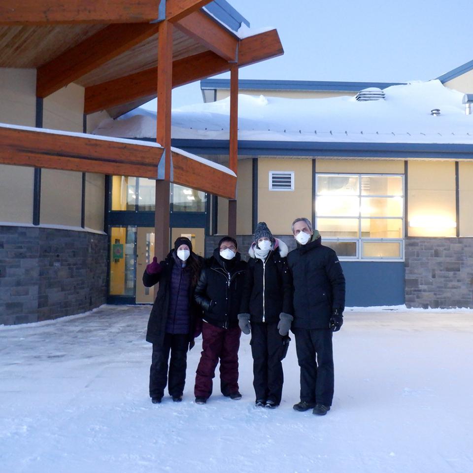 Four people are standing outside a building with winter coats, scarfs, gloves, and face masks on