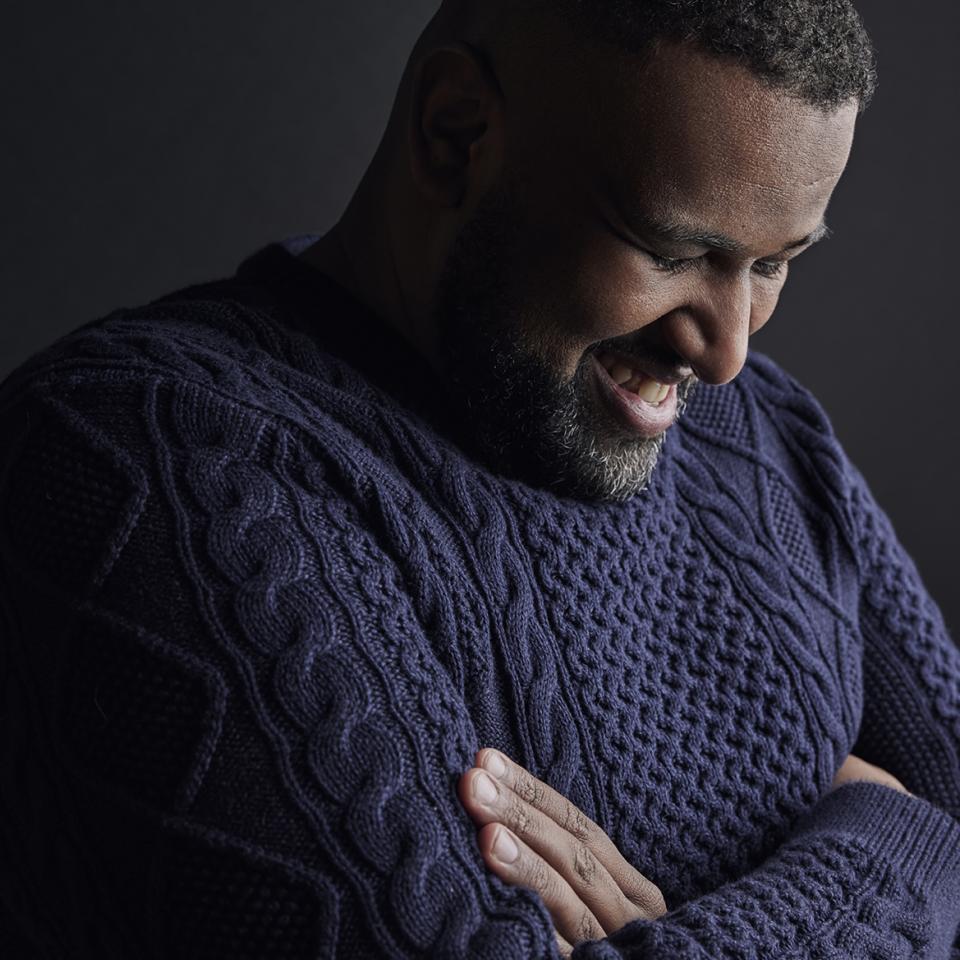 Elamin Abdelmahmoud, wearing a black knit sweater, stands against a black background, facing to his left, with his arms crossed, looking down and laughing. 