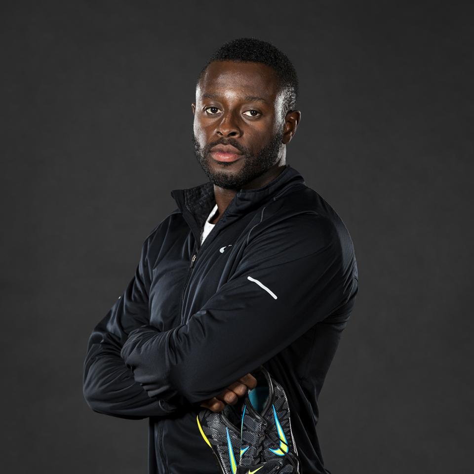 Sam Effah, arms crossed against his chest, holding a pair of running shoes, against a black backdrop.