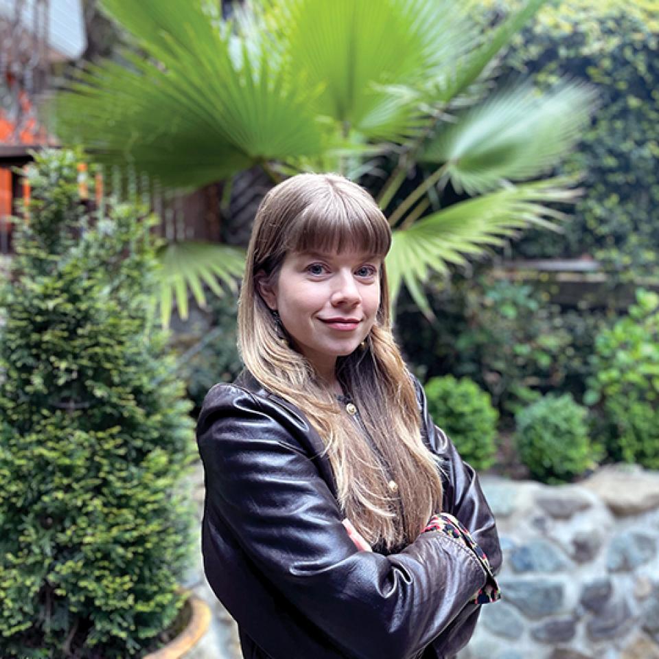 Young woman with long blond hair, wearing a leather jacket, stands in front of a garden.