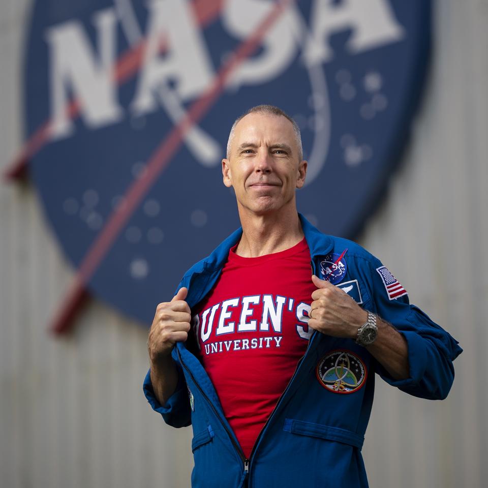 A man holds his zippered down NASA jumpsuit open to reveal a Queen's University T-shirt underneath.