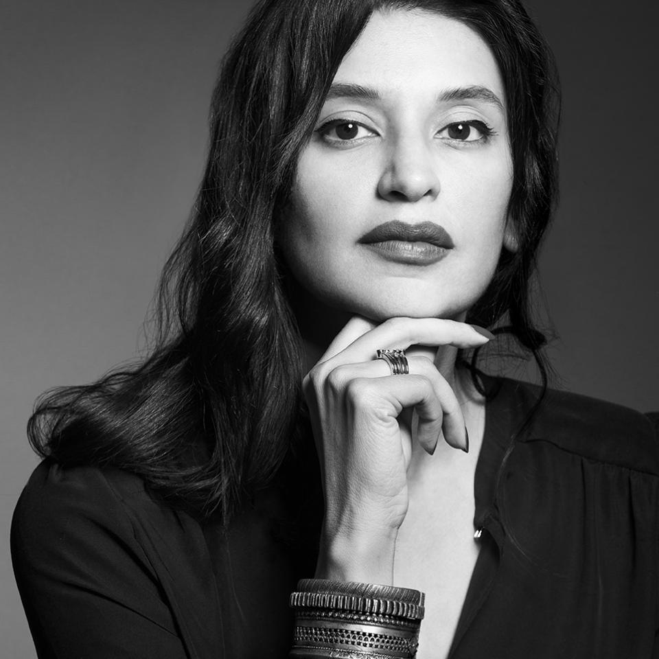 A woman with her left arm crossing in front of her and her right elbow on a table. Her chin rests on her right hand.