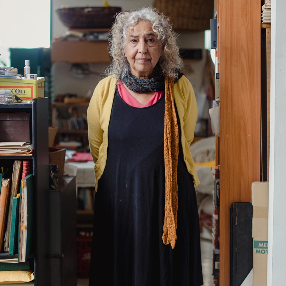 Woman standing in her art studio. She is looking straight ahead and surround by bookcases, art supplies, and portfolios.