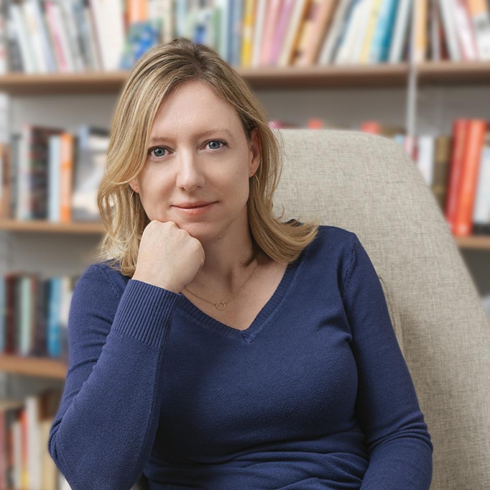 Woman poses for photo in a chair. One hand is in her lap and the other has her elbow on the arm of the chait and her fist resting on her chin.