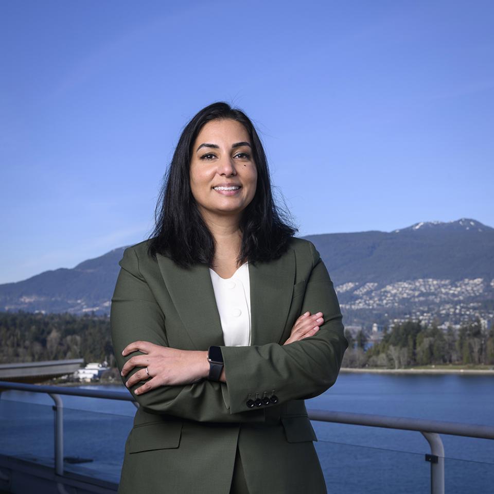 Niki Sharma stands with her arms crossed in front of water, mountains, and an open sky.