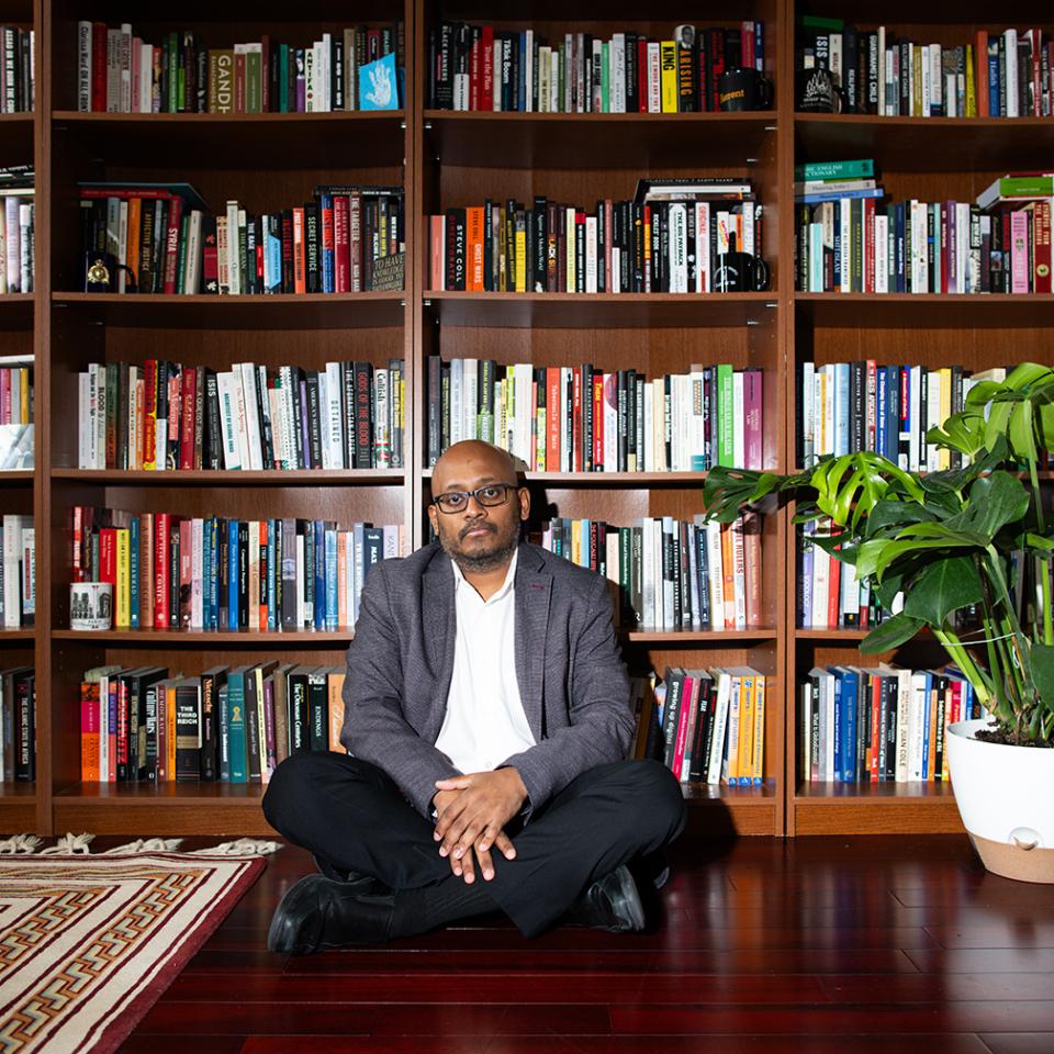 Amarnath Amarasingam sits cross-legged on the floor in front of a wall of bookcases. Beside him is a large leafy potted plant.