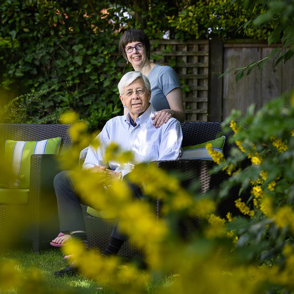 An older man sits on a wicker patio chair while a woman stands behind him, smiling with her hands on his shoulders. They’re surrounded by lush greenery and yellow flowers.
