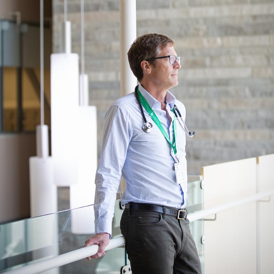 A man wearing glasses and a stethoscope stands by a glass railing, gazing thoughtfully into the distance. He wears a light blue shirt and green lanyard in a modern, well-lit building.