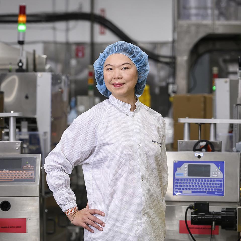 A woman wearing a lab coat and blue hairnet stands confidently in a manufacturing facility, surrounded by industrial equipment and machinery with indicator lights glowing.