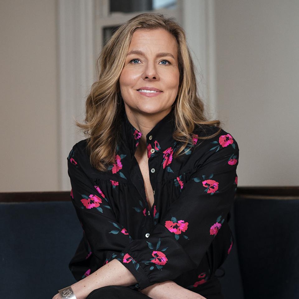 A woman with wavy blonde hair sits on a dark blue couch, smiling gently. She wears a black blouse with pink floral prints and a silver bracelet.