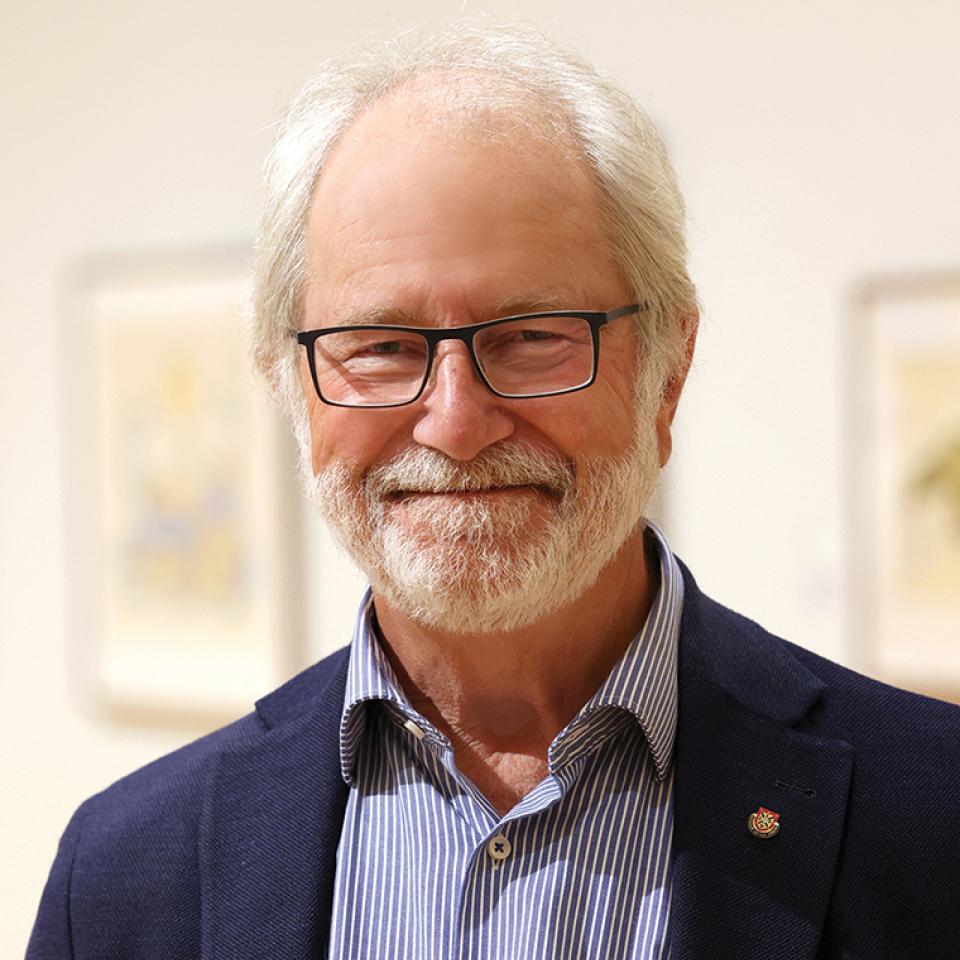A smiling older man with white hair, a beard, and glasses stands indoors wearing a navy blazer over a blue striped shirt, with framed artwork blurred in the background.