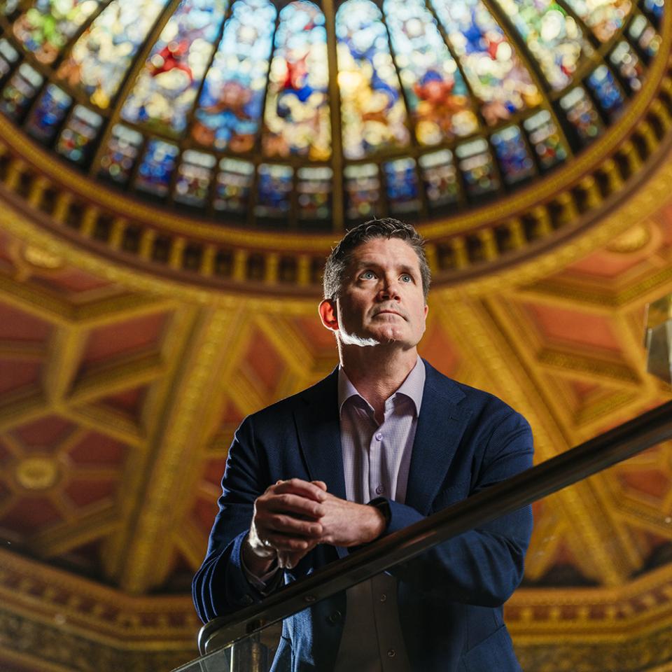 Portrait of a man in a suit standing beneath a stained-glass dome, looking upward with hands clasped on a railing.