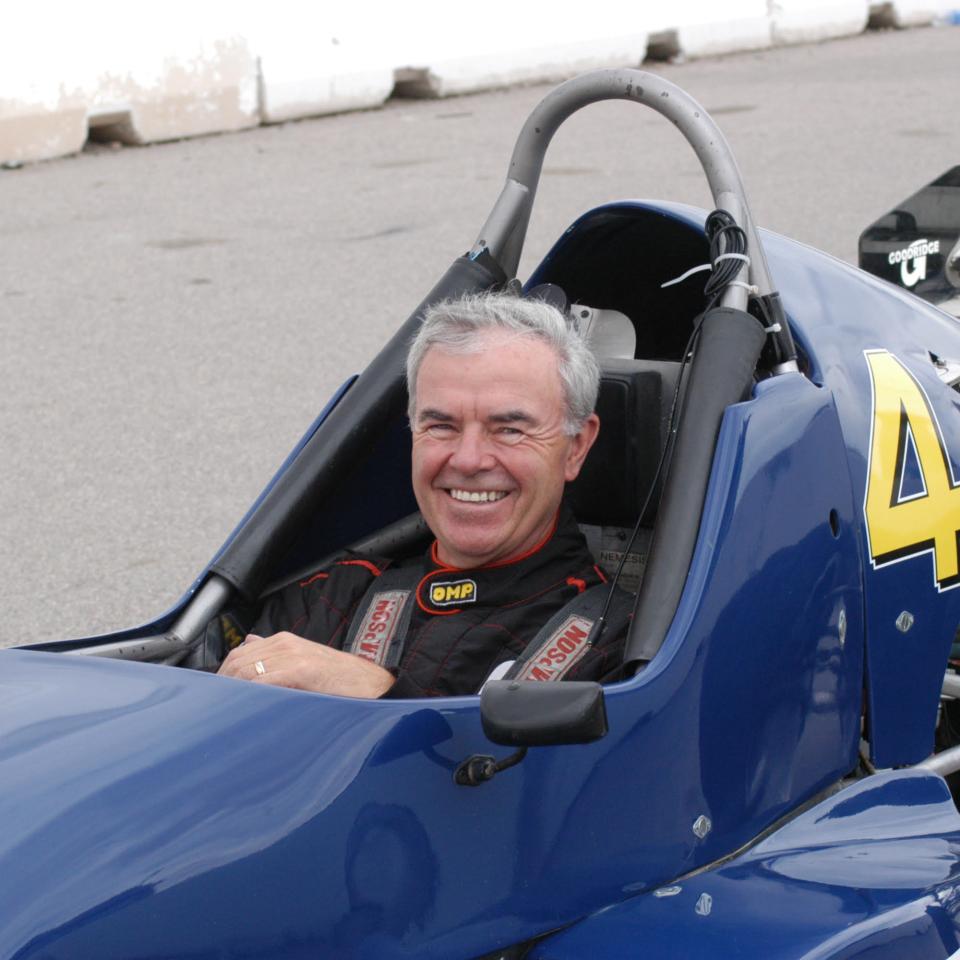 Man in a racing suit smiles while seated in a blue open-wheel race car marked with the number 4.