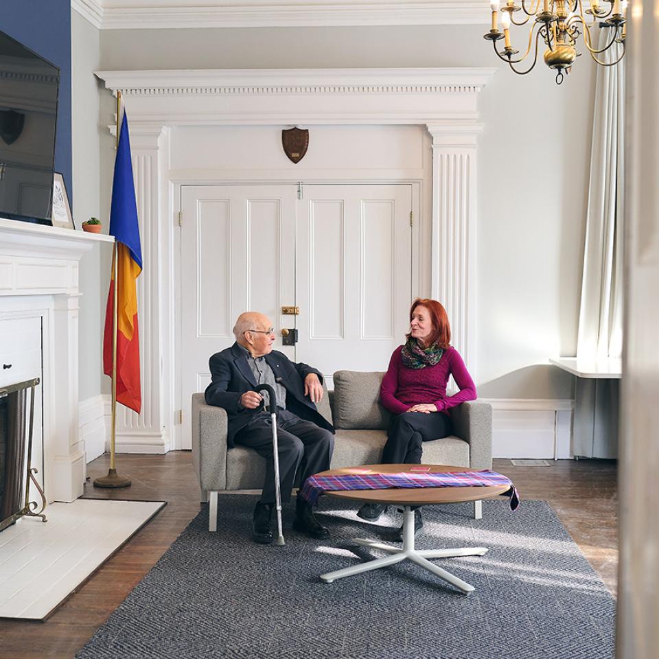Duncan Sinclair and Julie Epplett sit talking on a couch in a sunlit room.