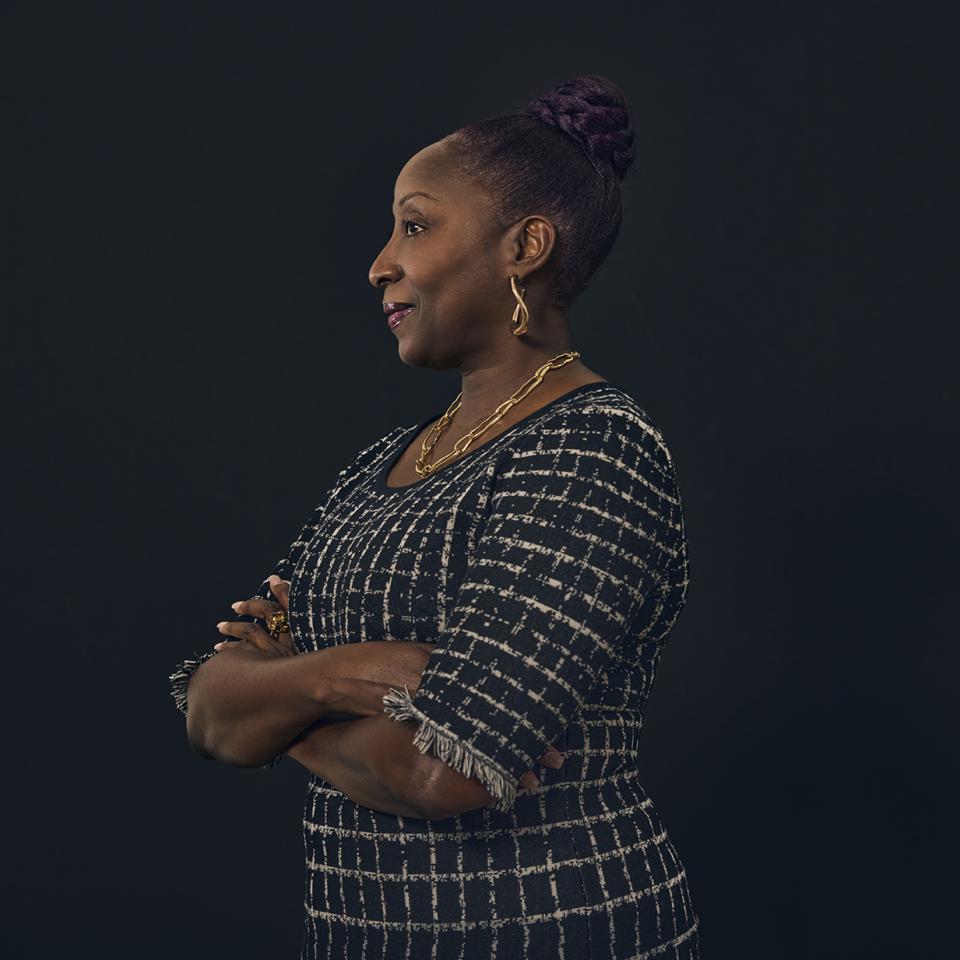 Profile portrait of a woman with arms crossed, wearing a patterned dress and gold jewelry against a dark background.