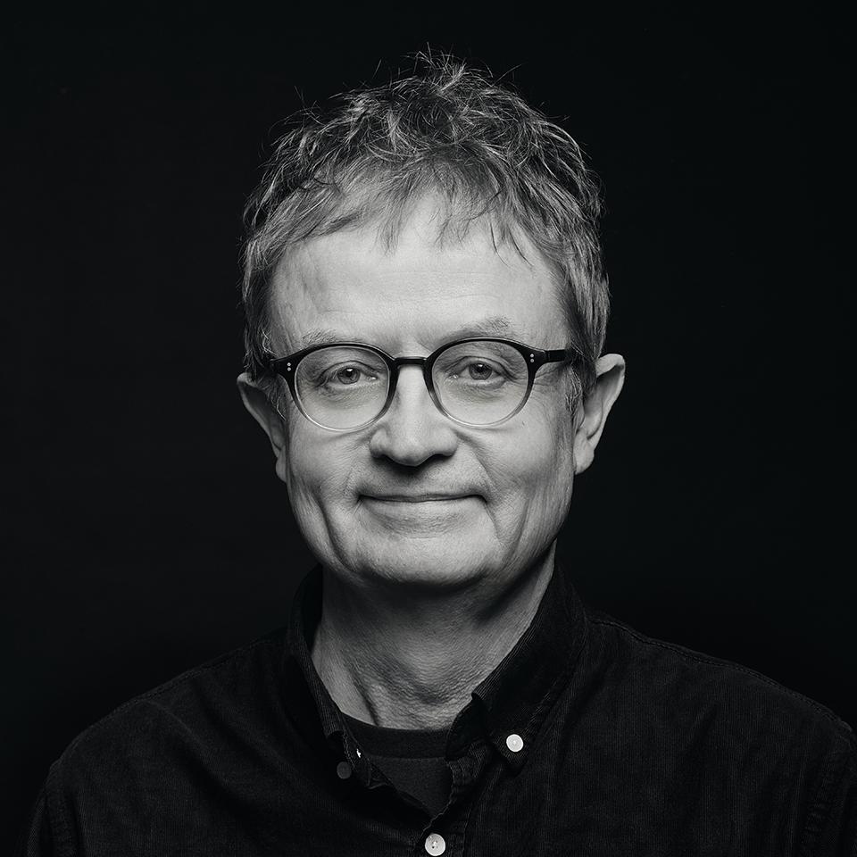 Black-and-white headshot of a man wearing glasses and a dark shirt against a black background.