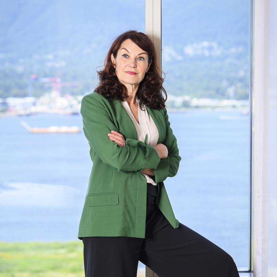 Woman in a green blazer posing beside a large window overlooking the water.