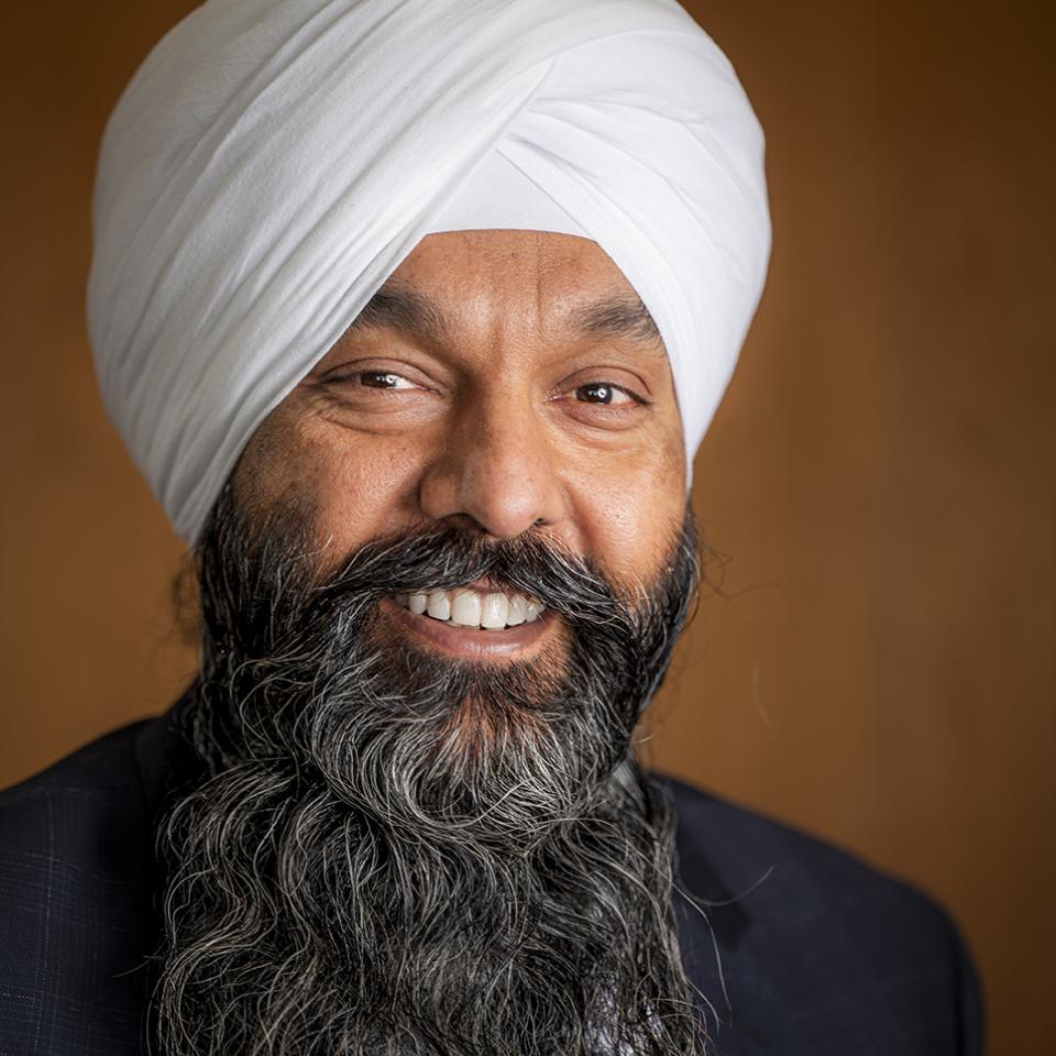 Smiling man wearing a white turban and dark suit against a warm brown background.