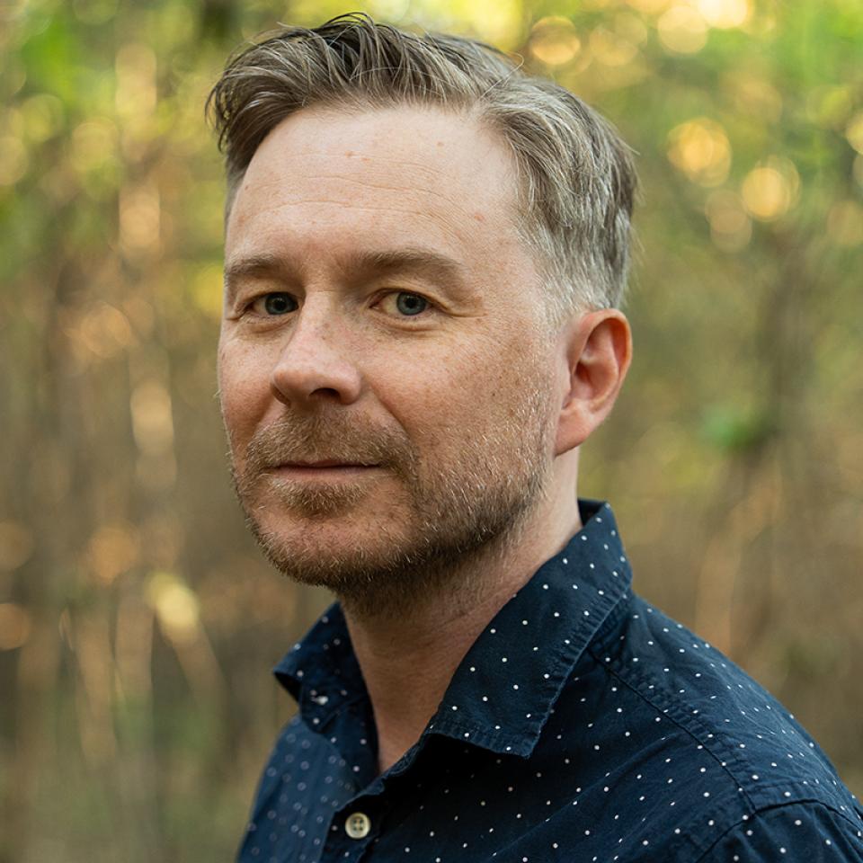 Man in a dark patterned shirt photographed outdoors in a wooded setting.