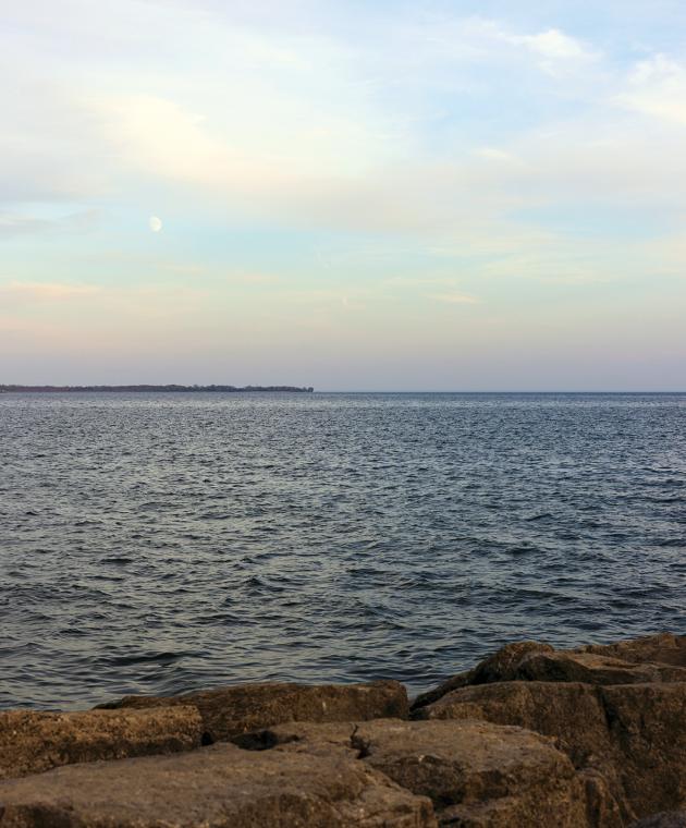 Lake Ontario with an early moon in the sky and rocky shoreline in the front.