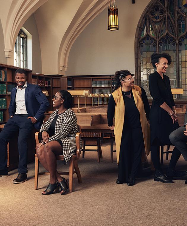 Eight members of the Black Studies faculty talk to each other in a group in a library. The library features a vaulted ceiling, arches, and stained glass windows.