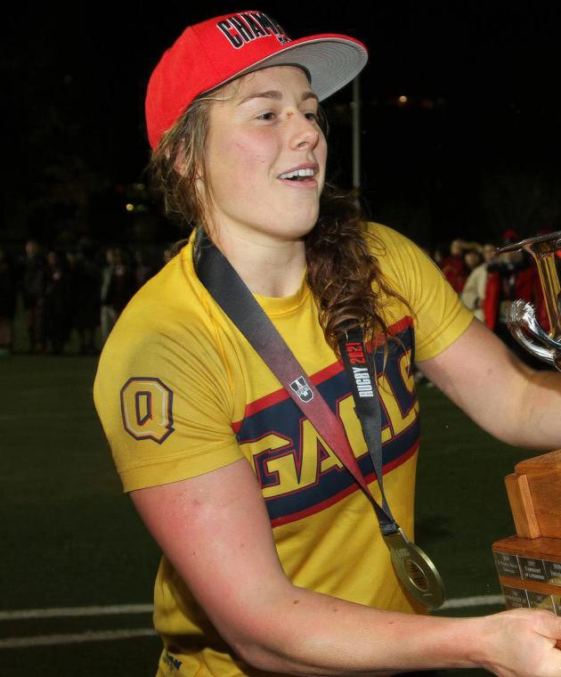Sophie de Goede is wearing a Queen’s Gaels jersey and red championship cap holds a large trophy on a field at night, celebrating a rugby victory.