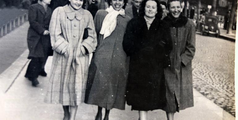 A old black and white stained photograph of Elizabeth Kawaley and three friends. They are wearing winter coats and walking towards the camera.