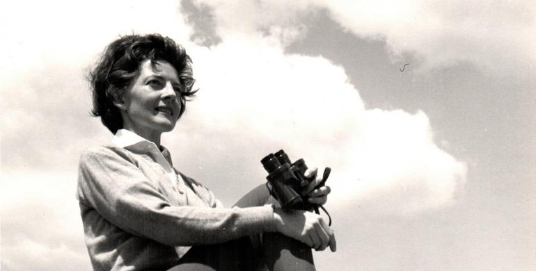 A photo from the 1960s shows a woman sitting on a large rock. Her right leg is pulled up towards her body and her hands are holding her knee. In her left hand is a set of binoculars.