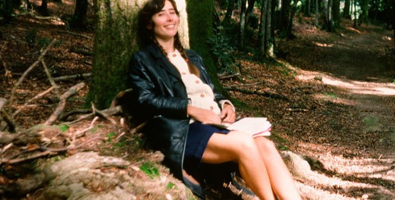 An older photo of a yong Connie Eaves sitting under a tree in a forrest.