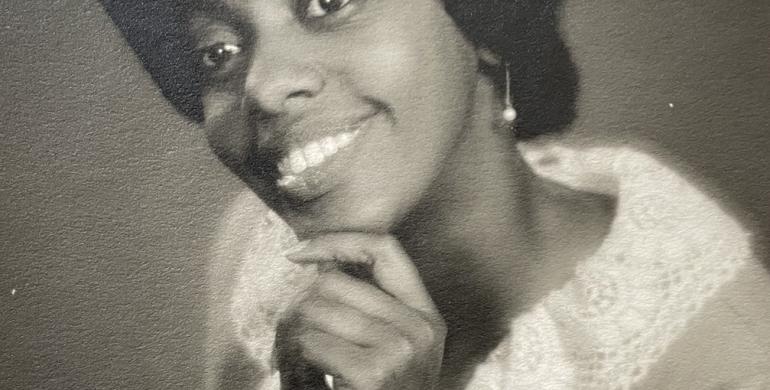 A vintage black-and-white portrait of a woman smiling warmly, resting her chin on her hand. She wears a dress with lace trim and pearl earrings, posed gracefully.