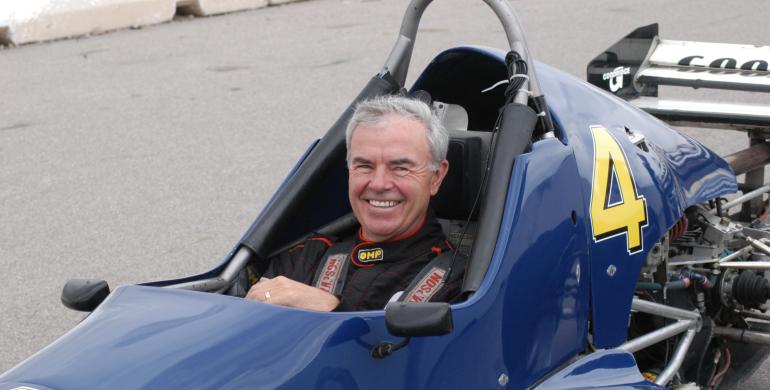 Man in a racing suit smiles while seated in a blue open-wheel race car marked with the number 4.