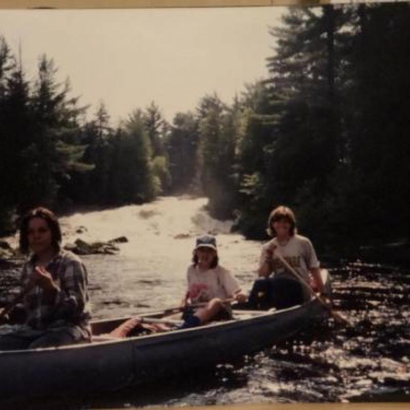 Three people in a canoe. A lake lined with evergreen trees in the background.