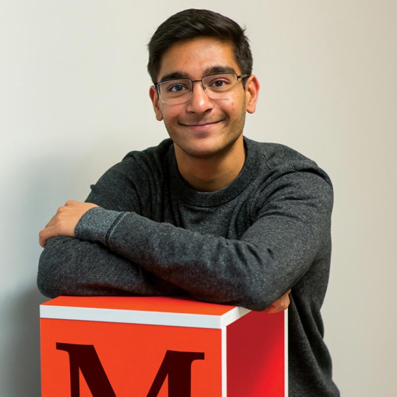 Rahul Patel, Artsci'21, sitting on a bench in the lab, leaning on a large block with the letter M on it.