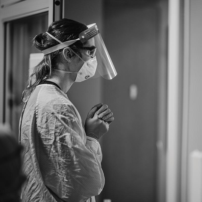 An ICU nurse standing in a doorway wearing a gown, gloves, mask, and facial shield.