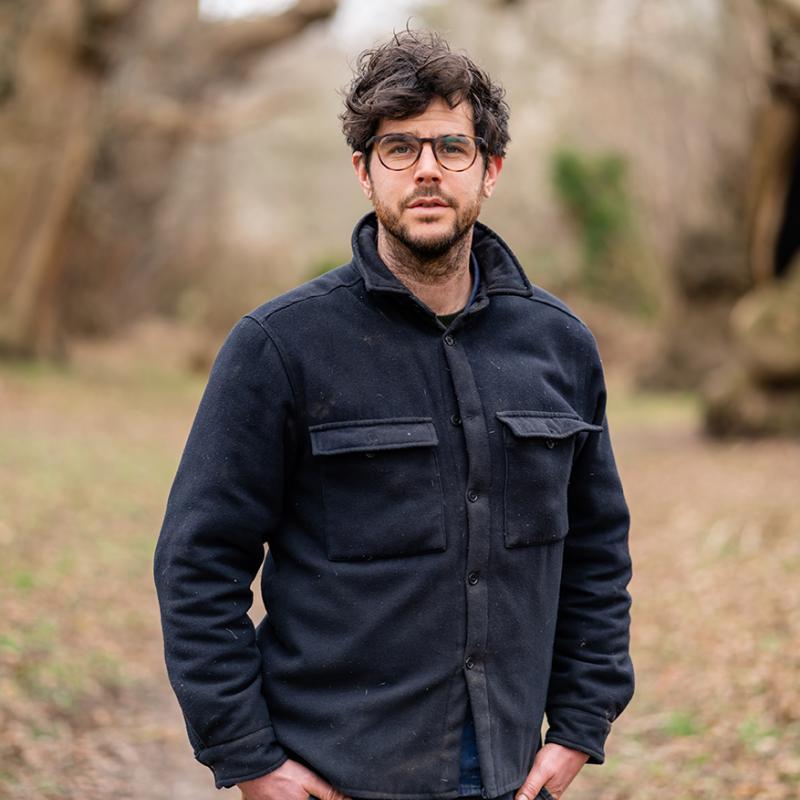 Guy Lucas standing in the woods wearing a black shirt jacket and dark-rimmed glasses