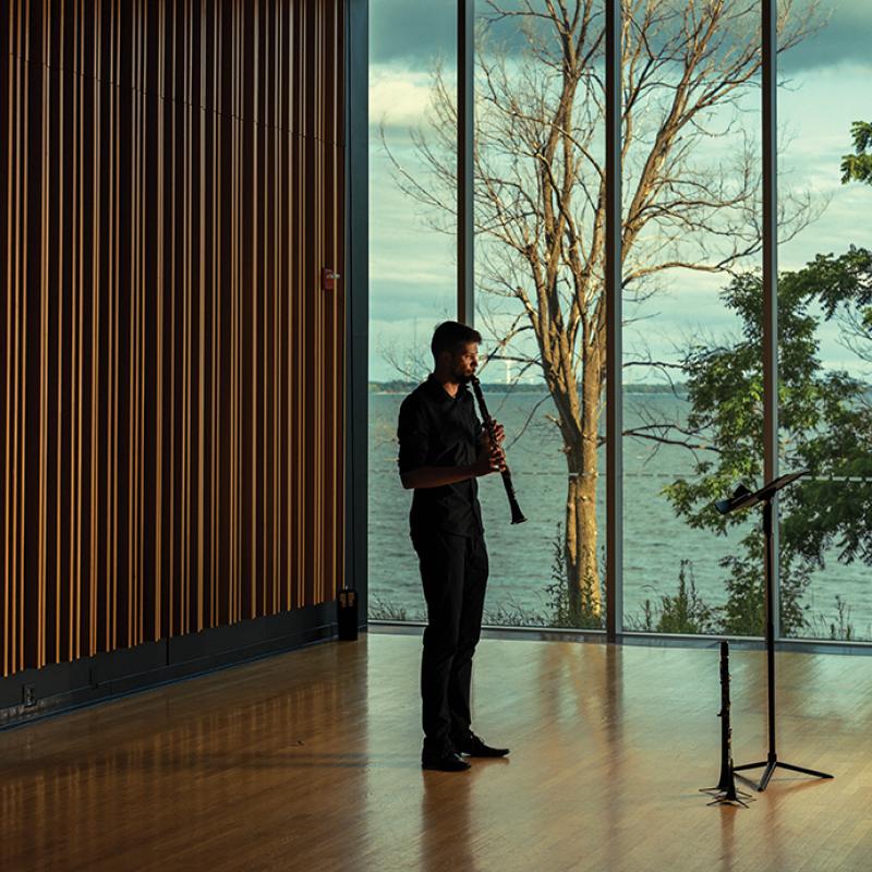 Man playing clarinet in front of glass window overlooking the water.