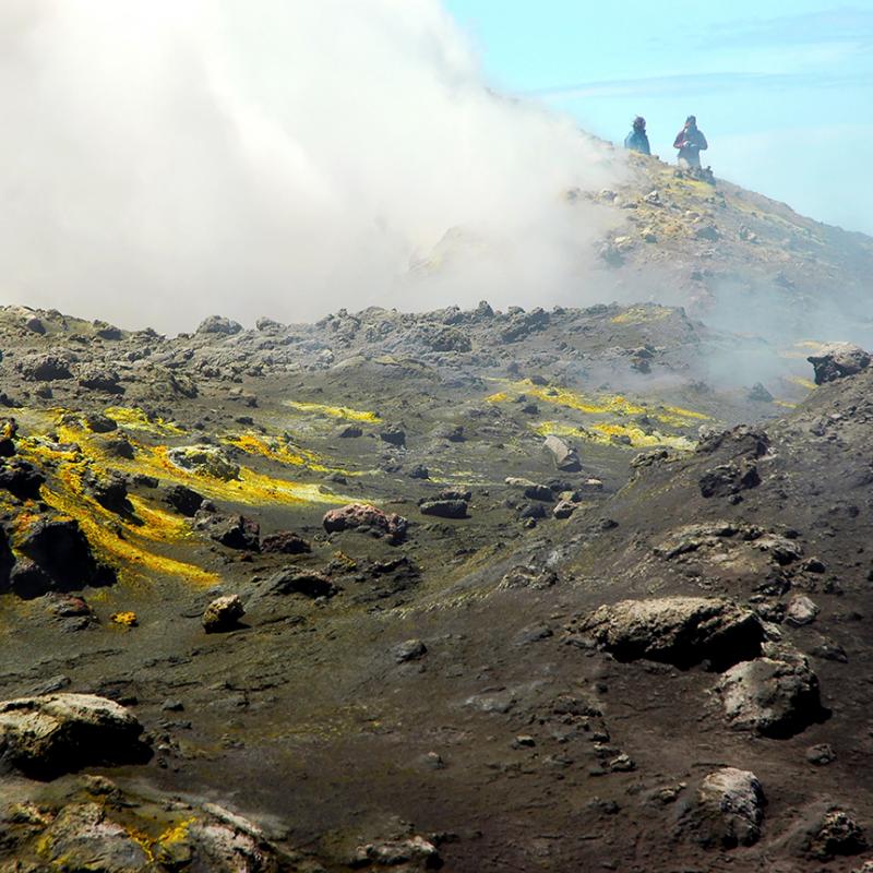 Three people walk atop a rocky and smoky Mount Etna