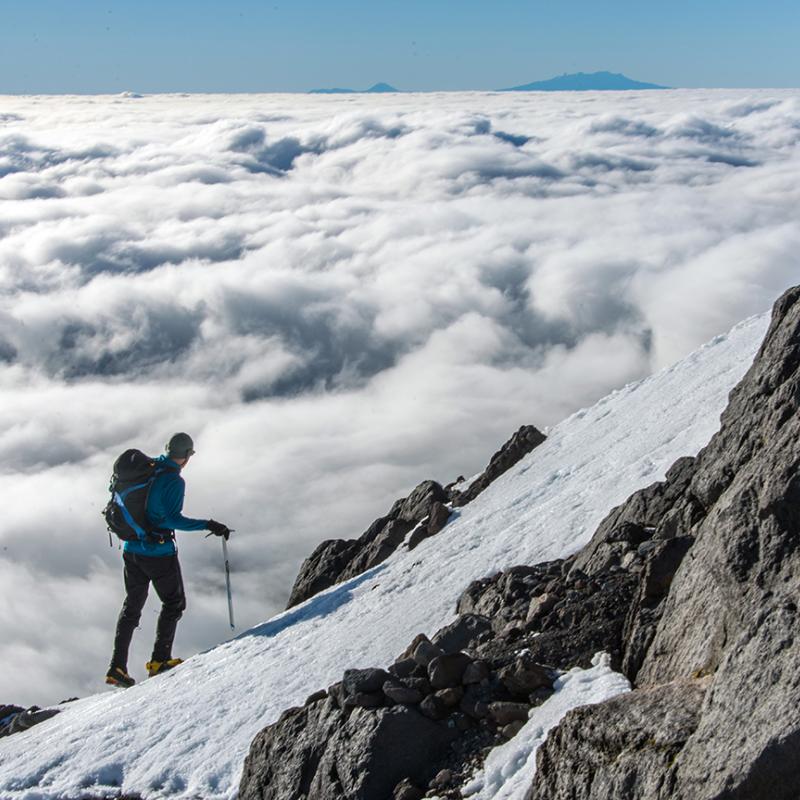 A man stands on a snowy mountainside looking out at the clouds below him