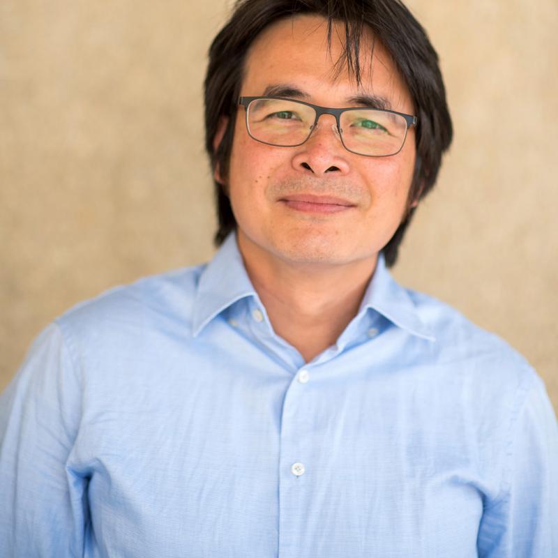 Asian man wearing black-rimmed glasses and a blue shirt against a beige wall.