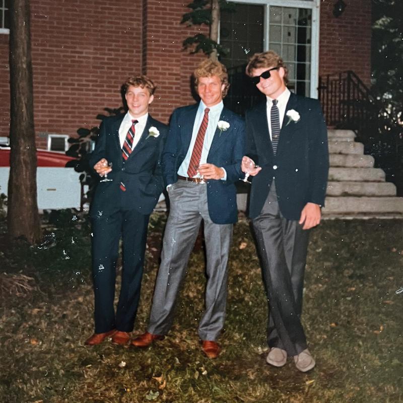 Four young men dressed in suits stand in front of a brick house.