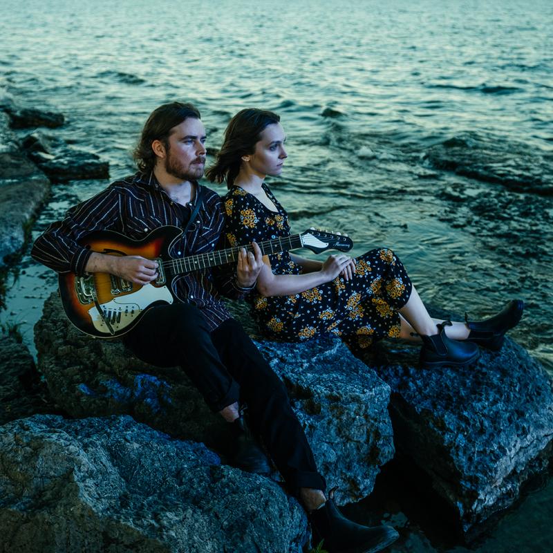 A man, playing a guitar, and a woman, leaning against him, look out at the lake as they sit on large rocks by the shoreline.