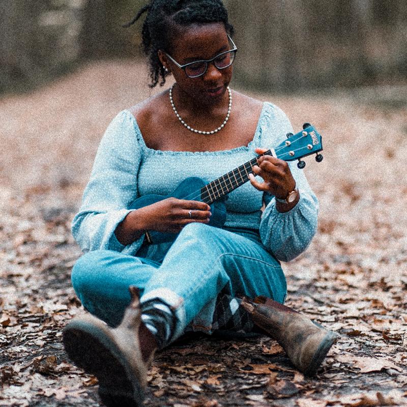 A woman plays a ukulele while sitting on the leaf-covered grounds of a forest.