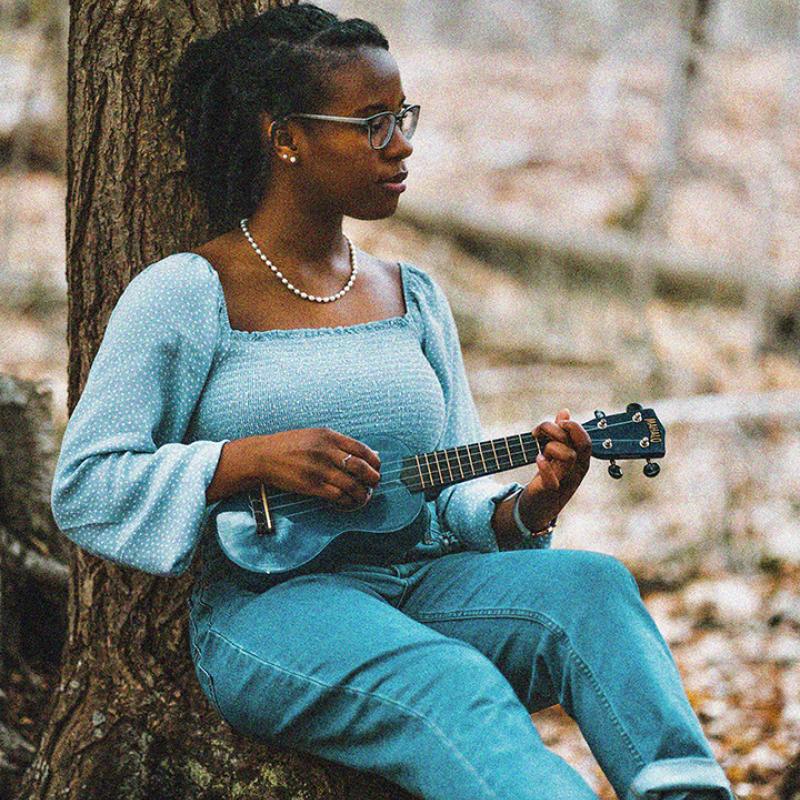 A woman leaning against a tree in the woods playing a ukulele.