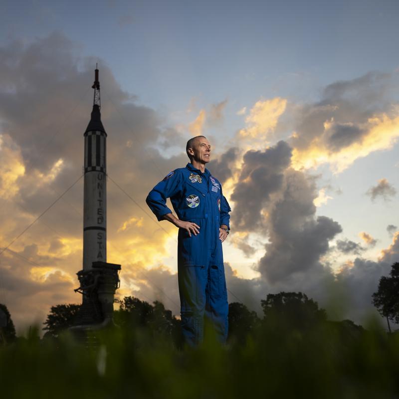 Astronaut, Drew Feustel, stands in his astronaut jumpsuit, in front of two rockets and a cloud-filled sky behind them.