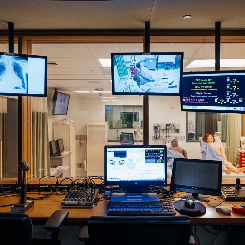 View inside a room with multiple monitors that are displaying medical information related to the two SimMan 3G Plus' that are seen through a window.