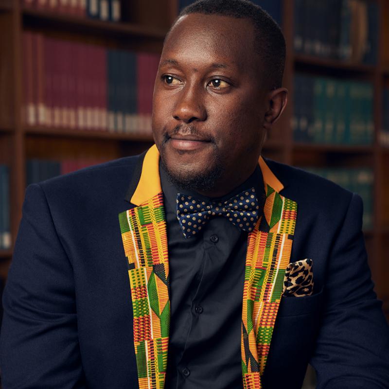 Dalitso Ruwe sits at a table with a wall of library books in the background.
