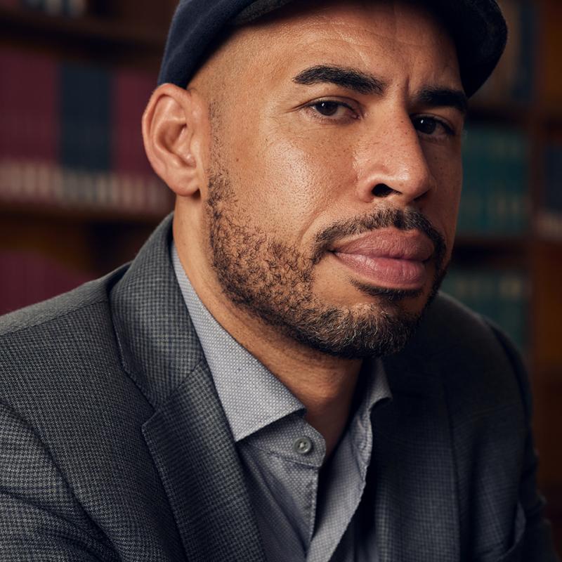 Daniel McNeil sits in front of a wall of books. He is wearing a news cap and suit jacket.