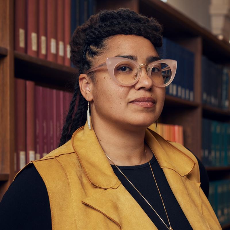 Jennifer Leath stands in front of a wall of books. She is wearing cats eye glass, feather earrings, and a long necklace.